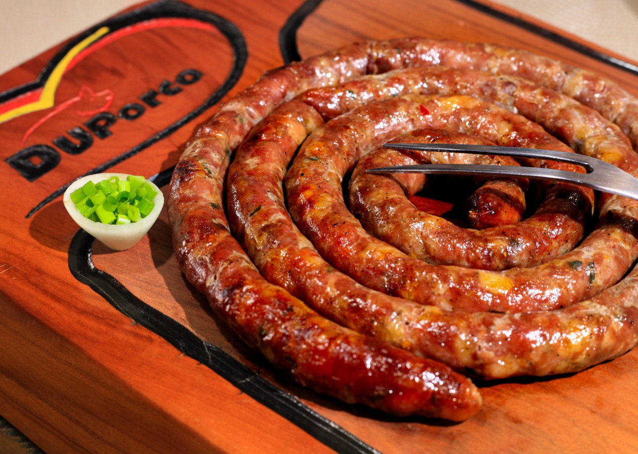 Linguiça Mineira DuPorco grelhada em espiral, servida com garfo sobre tábua com a marca DuPorco